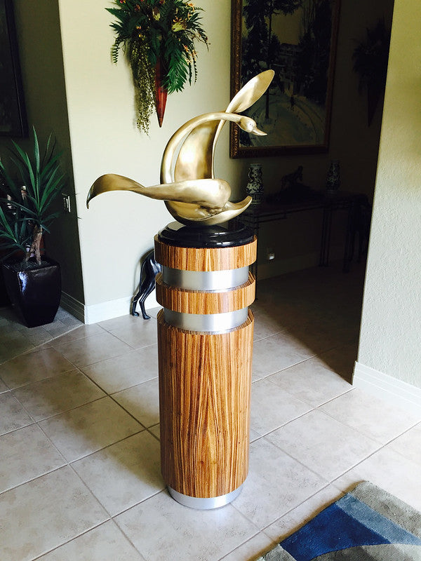 wood grain pedestal with two silver bands wrapped around and a matching silver toe kick. There is an abstract bronze/gold statue on top.