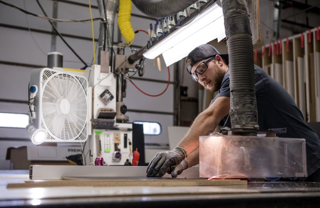 man wearing safety glasses and hat cutting wood on a band saw. there is a white box fan in th eleft corner