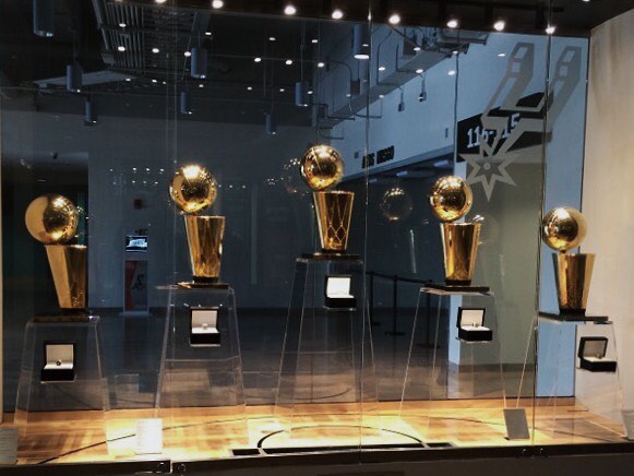 There are five clear acrylic trapezoid prisms lined up against a reflective wall. The center is the tallest, with the pedestals on each side getting shorter to create a pyramid effect. The prisms are pedestals holding golden Spurs Championship trophies. The trophies are gold cylindrical shapes with golden globes perched on the top. 