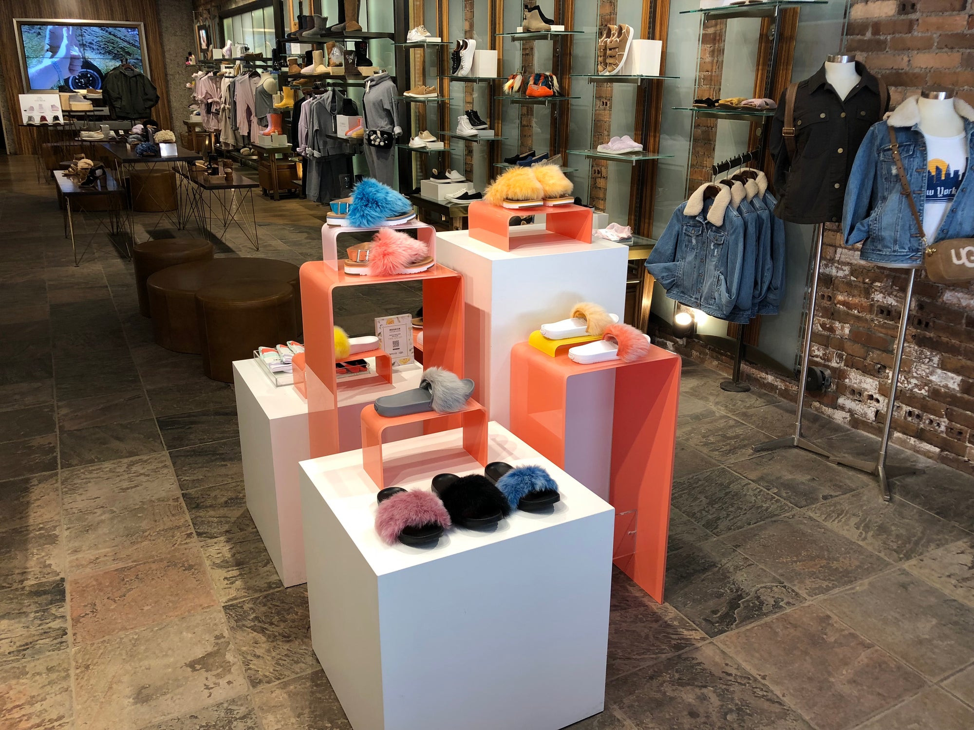  Photo is set in retail space/ clothing store. The focus is on the pedestal display. there are two white cube pedestal and one taller rectangular pedestal. There two tall waterfall pedestals that are a pink coral color. There are two smaller versions of the coral pedestals on top of the tall rectangle pedestal and the white cube in the front. There are fuzzy slippers on the displays.   