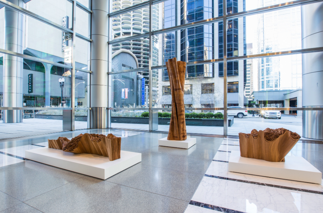 Museum floor with giant windows. There are three large white pedestals that are low to the ground. They are displaying wooden sculptures reminiscent of trees. 