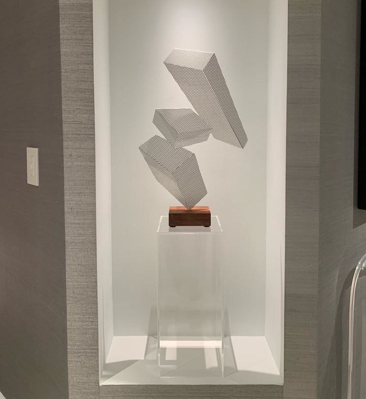clear acrylic pedestal with modern cube art on top. it looks like three rectangular cubes made of mesh or wire are precariously and almost impossibly balanced on a small wooden step up. 