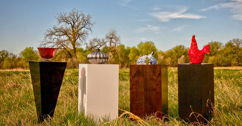 Four pedestals outside in a grassy field. From left to right: black laminate tapered pedestal with glass red bowl. White rectangular pedestal with silver orb-like piece on it. dark wooden grain rectangle pedestal with two ceramic white rabbits. a darker wood pedestal with a bright red chicken sculpture.